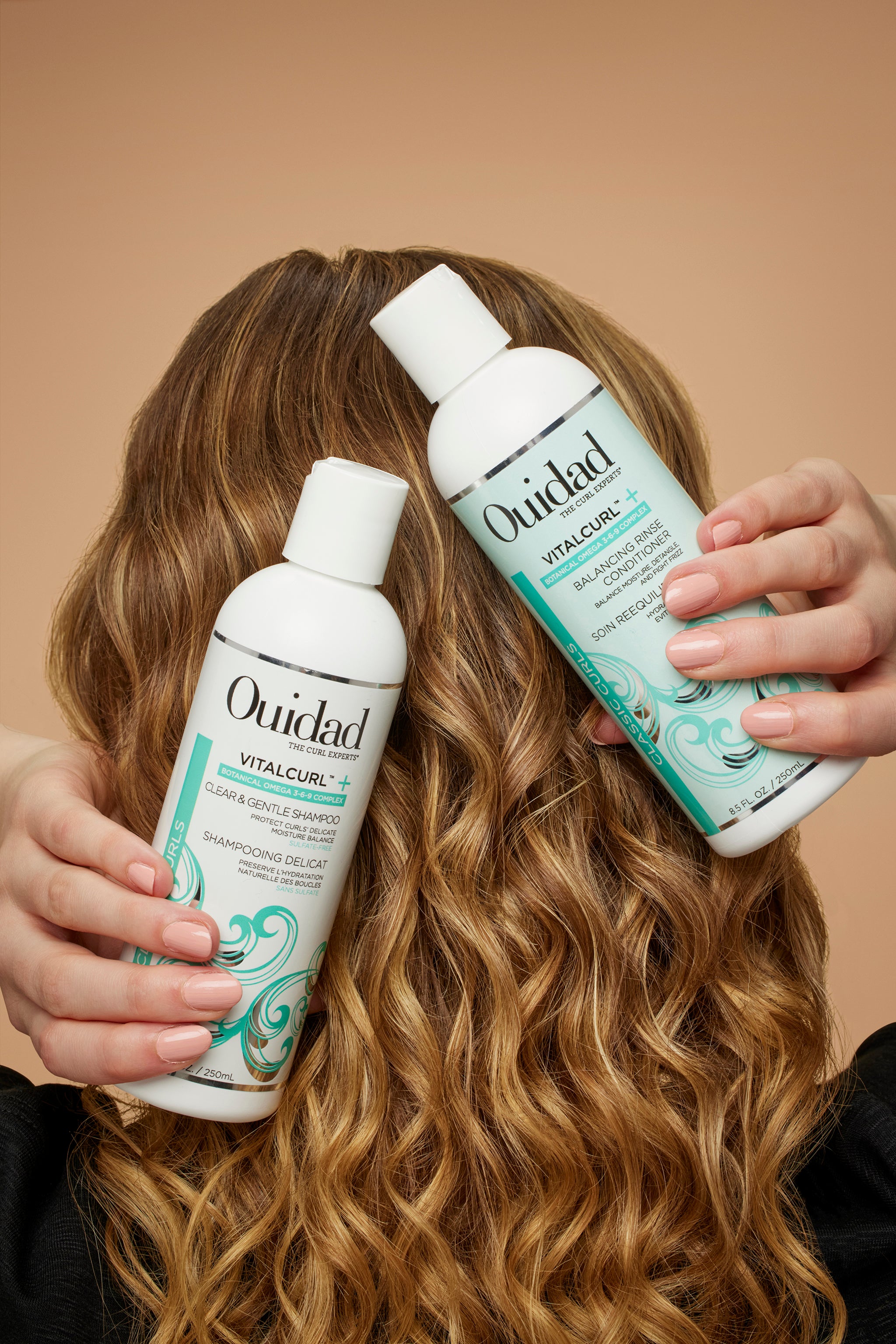 Curly haired model holding bottles of VitalCurl®+ Balancing Rinse Shampoo and Conditioner